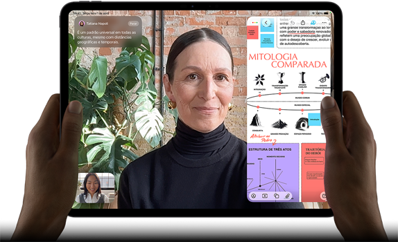 Parte da frente do iPad Pro preto-espacial na horizontal, cantos arredondados, tela com borda preta, mãos segurando os lados esquerdo e direito, tela mostrando uma conversa no FaceTime sendo traduzida ao vivo e um app de educação com uma apresentação sobre "Mitologia Comparada" com textos, diagramas e anotações manuscritas