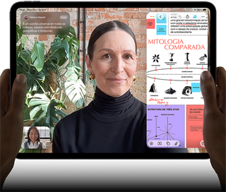 Parte da frente do iPad Pro preto-espacial na horizontal, cantos arredondados, tela com borda preta, mãos segurando os lados esquerdo e direito, tela mostrando uma conversa no FaceTime sendo traduzida ao vivo e um app de educação com uma apresentação sobre "Mitologia Comparada" com textos, diagramas e anotações manuscritas
