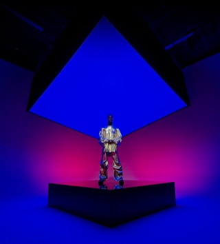 Imagem de uma pessoa vestida com uma roupa metálica em pé em um palco quadrado que se estende até o fundo, capturada na câmera ultra-angular do iPhone 16 Pro Max