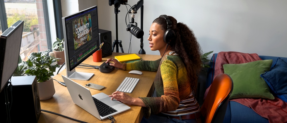 Uma mulher em casa usando fones de ouvido e falando no microfone trabalha na mesa com o MacBook Pro conectado a dois monitores externos