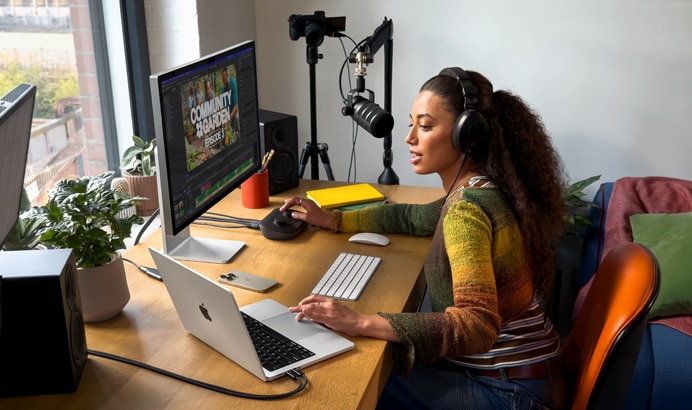 Uma mulher em casa usando fones de ouvido e falando no microfone trabalha na mesa com o MacBook Pro conectado a dois monitores externos