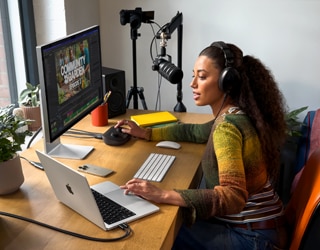Uma mulher em casa usando fones de ouvido e falando no microfone trabalha na mesa com o MacBook Pro conectado a dois monitores externos