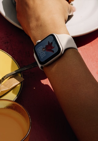 Braço de uma pessoa segurando o café da manhã, Apple Watch SE 3 no pulso, caixa estelar de alumínio, tela Sempre Ativa, retrato de uma pessoa no mostrador, pulseira esportiva estelar